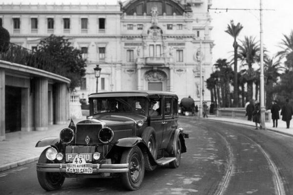 Zwart-witfoto van een klassieke rallyauto met het bord &lsquo;Rallye Automobile Monte-Carlo&rsquo; aan de voorzijde, rijdend over een brede straat in Monte Carlo. Op de achtergrond staat een monumentaal gebouw, omgeven door palmbomen en enkele voetgangers.