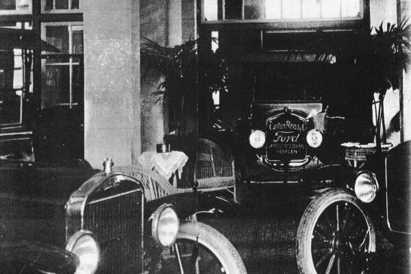 Historische zwart-witfoto van een showroom of garage met meerdere vroege automobielen. Op de voorgrond staat een open personenauto met grote koplampen en smalle spaakwielen. Daarachter is een Ford opgesteld met op de voorzijde het opschrift &ldquo;Canton Reiss & C. &ndash; Ford &ndash; Official Dealer &ndash; Heerlen&rdquo;. De auto&rsquo;s staan tussen planten en meubilair in een overdekte bedrijfsruimte.