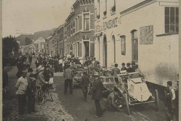 Historische zwart-witfoto van deelnemers aan de rally Parijs-Amsterdam-Parijs tijdens een rustmoment onderweg naar de grens bij Eijsden. In een met kinderkopjes geplaveide straat staan meerdere vroege open automobielen achter elkaar opgesteld, voorzien van startnummers. Bestuurders, begeleiders en omstanders verzamelen zich rond de voertuigen, terwijl langs de straat een grote groep belangstellenden toekijkt. Op de achtergrond zijn karakteristieke woon- en bedrijfsgebouwen zichtbaar.